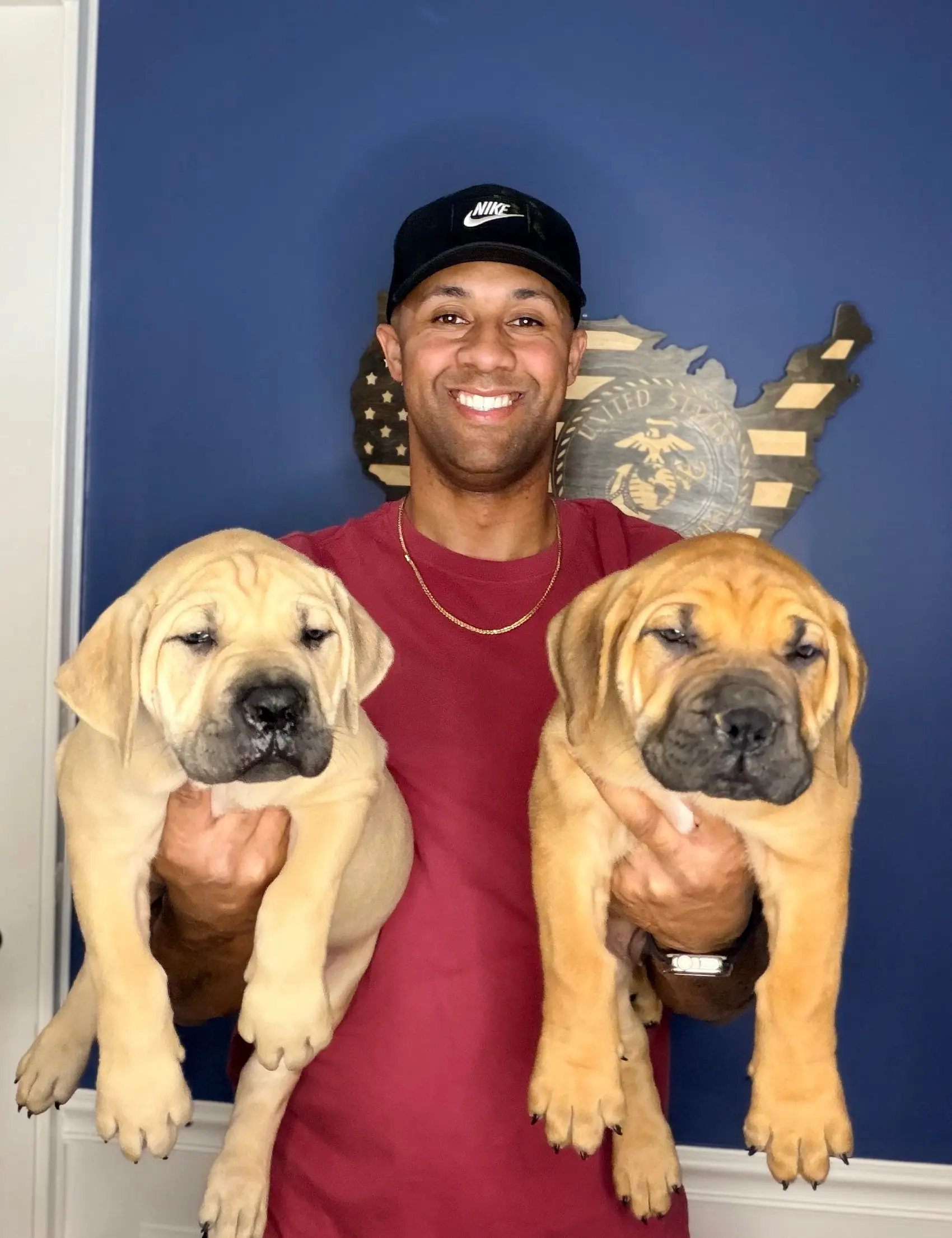 The veteran owner of LowCountry Boerboels holding two South African Boerboel puppies, one in each arm, under a wooden US Marine Corps flag